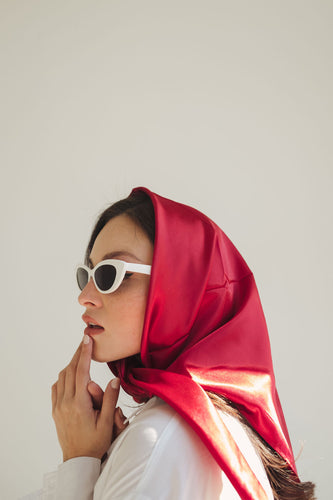 Side profile of a white woman with white sunglasses touching her lip. She is wearing a ruby red scarf around her head and neck, revealing some of her long brown hair.