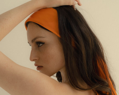 Side profile of a white woman with green eyes and brown hair wearing a bright orange satin scarf as a headband