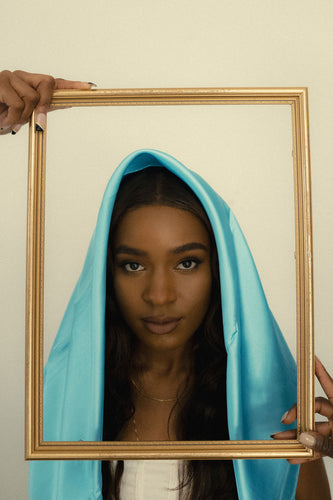 A black woman with brown hair wearing a cyan satin scarf on top of her head looks into the camera. Two hands on either side of her hold a gold picture frame around her face. 