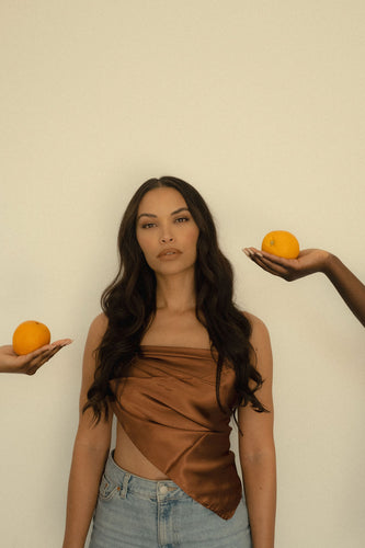 Olive-skinned woman with rib-length brown hair wears a chocolate brown satin scarf as an asymmetrical triangle top. Two hands hold oranges on either side of her.