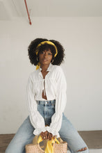 Load image into Gallery viewer, Black woman sits on top of a stool, wearing a white ruffle shirt and jeans. She has a bright yellow scarf as a headband in her curly afro and is holding a wicker bag that has a bright yellow scarf tied around its handle.
