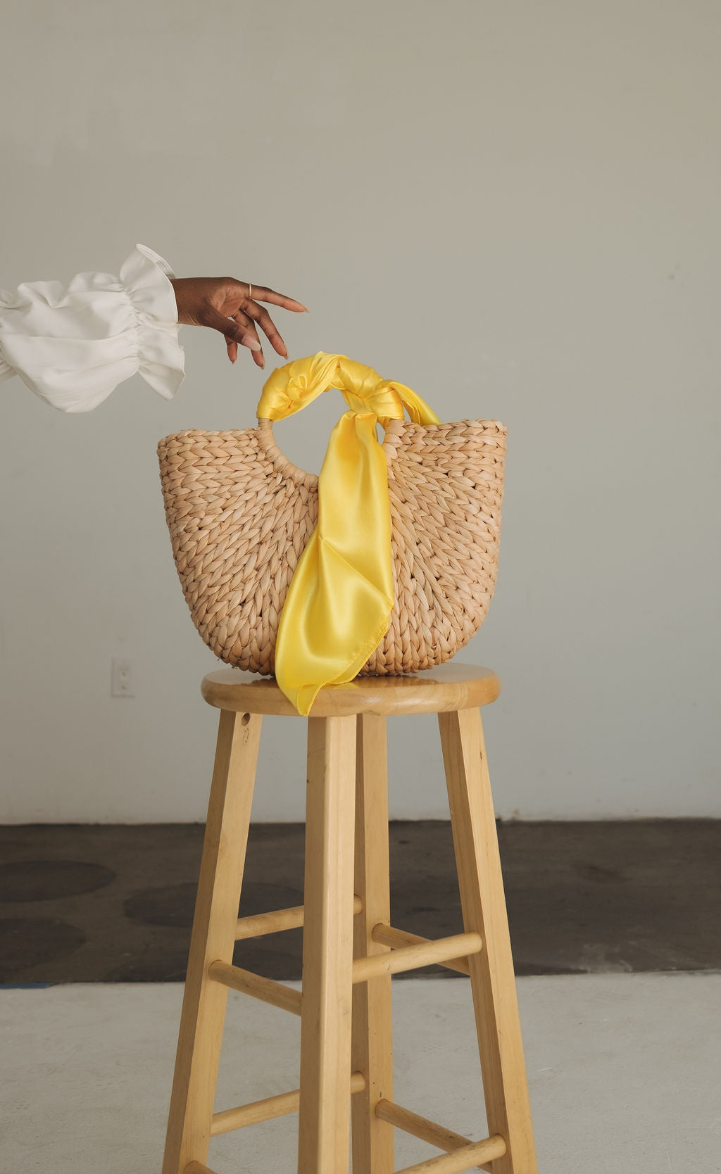 A tan wicker bag sits atop a stool. A woman's hand is reaching for the handle, which is wrapped in a bright yellow satin scarf.