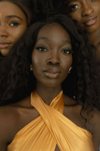 Load image into Gallery viewer, Dora Owusu looks into the camera, wearing a gold satin scarf as a criss-cross halter top. Two Black women look into the camera behind her.
