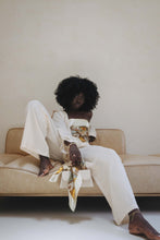 Load image into Gallery viewer, Darkskinned Black woman with an afro leans against the back of a beige couch in front of a beige wall, with her right foot rested on the couch. She is wearing loose ivory pants and an open ivory top, exposing the patterned, ivory satin scarf she is wearing as a top. In her right hand, she holds a white purse that has the same scarf tied around the handle. 
