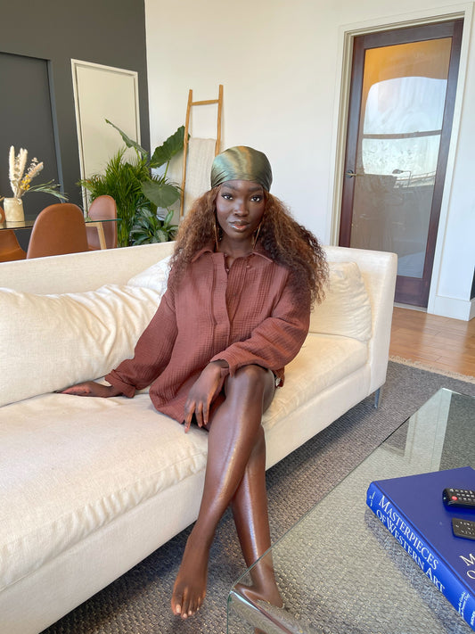 Dora Owusu sits cross-legged in a brick-colored button down on a white couch, wearing a khaki green satin scarf as a head scarf