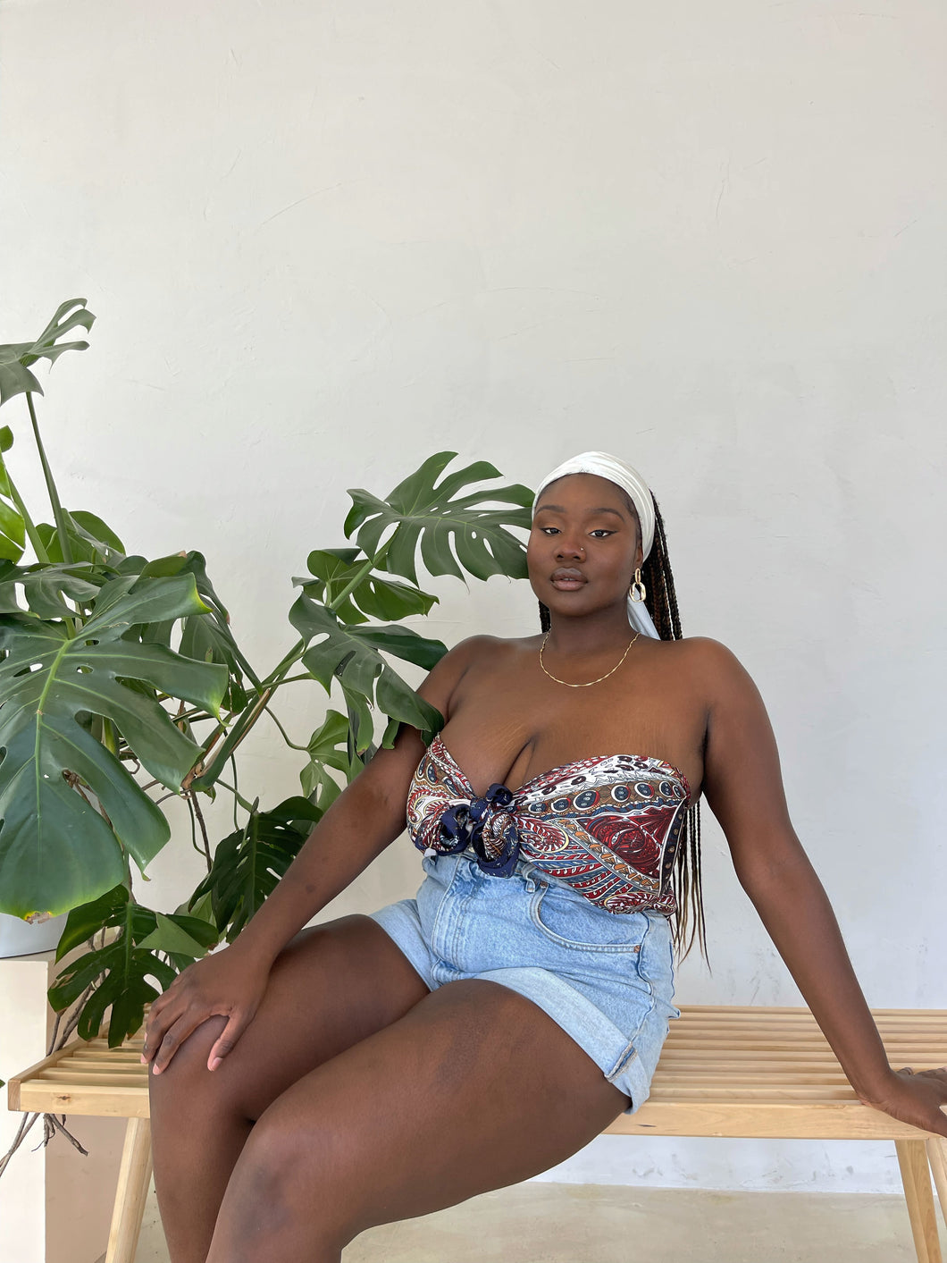 A Black woman sits on a bench, leaning on her left arm, in front of a white wall. She sits to the right of a monstera plant. She is looking directly at the camera while wearing denim shorts, an ivory scarf as a headband, and a red, white, and blue feather and paisley patterned scarf as a twist-front top.