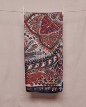 Load image into Gallery viewer, Rectangular, folded, ivory satin scarf with red, white, and blue feathers and paisley and floral accents is held on a clothesline by two clothespins in front of a light beige background
