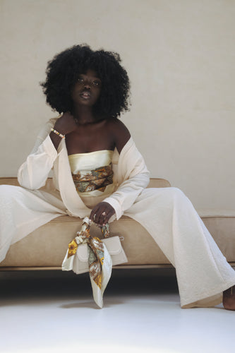 Darkskinned Black woman with an afro sits on a beige couch in front of a beige wall. She is wearing loose ivory pants and an open ivory top, exposing the patterned, ivory satin scarf she is wearing as a top. In her left hand, she holds a white purse that has the same scarf tied around the handle. 